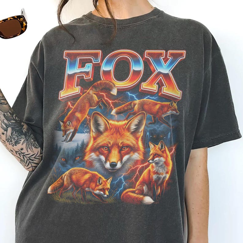 May include: Dark gray t-shirt featuring a vibrant graphic of foxes and the word "FOX" in a retro font. The design includes multiple foxes, lightning bolts, and a forest scene, creating a vintage, outdoorsy aesthetic.