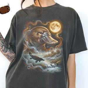 Puede incluir: Camiseta gris oscuro con una ilustración detallada de un ornitorrinco y una luna llena. El diseño incluye un ornitorrinco nadando en el agua, con una cascada y burbujas. La camiseta está hecha de un material suave.