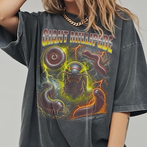 May include: A charcoal gray oversized t-shirt featuring a graphic print with the text GIANT MILLIPEDE in yellow and red. The design displays multiple brown millipedes surrounded by yellow lightning bolts and green energy effects on the front.