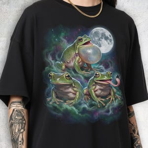 Three realistic green frogs positioned under a large luminous full moon. The top frog has an inflated translucent vocal sac as if croaking. Below, one frog faces forward with a calm expression, while the other sticks its long tongue out to catch a fly. Cosmic teal and purple nebula mist with stars in a retro 90s galaxy style.