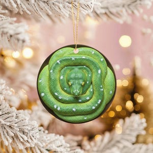 May include: A round Christmas ornament featuring a coiled green snake with white spots. The snake is centered on a black background. A gold string is attached for hanging. The ornament is set against a blurred background of a decorated Christmas tree.