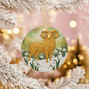 May include: A round Christmas ornament featuring a golden ram with large curved horns, standing in a snowy field with white flowers. The ram has detailed scrollwork. The background is a soft green and white. A gold string is attached for hanging.
