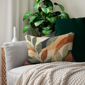 May include: Rectangular decorative pillow with a botanical design featuring green, brown, and beige leaves and abstract shapes. The pillow is on a gray sofa with a cream-colored throw blanket. A dark green pillow is behind it.