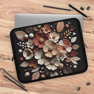 Puede incluir: Funda negra para portátil con un diseño floral en tonos crema, melocotón y marrón. Las flores y hojas están dispuestas en un patrón tridimensional detallado. La funda tiene una cremallera negra y está sobre una superficie de madera.