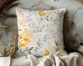 May include: Square decorative pillow with a botanical design. The pillow features a white background with yellow and gray floral and leaf patterns. The pillow is placed on a cream-colored surface.