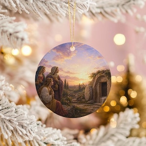 May include: Round ceramic Christmas ornament featuring a religious painting of three women at a tomb. The artwork depicts a sunrise with a warm color palette of pinks, oranges, and blues. A gold string is attached for hanging.