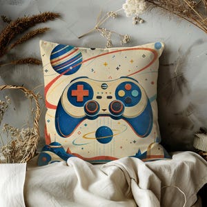 May include: A square throw pillow featuring a cream-colored background with a central illustration of a blue and beige video game controller. The design includes a striped planet, a smaller blue planet with rings, and small colorful plus signs.