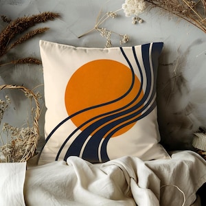 May include: Square decorative pillow with a cream-colored background. The pillow features an orange sun design with navy blue and cream-colored curved lines. The pillow is placed on a white sheet.