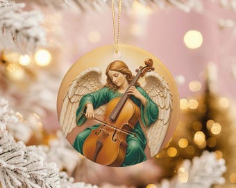 May include: Round Christmas ornament featuring an angel playing a cello. The angel has long auburn hair and wears a green robe. The cello is a warm brown color. The background is a soft yellow with white angel wings.