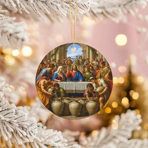 May include: Circular Christmas tree decoration displaying a classical painting of a religious scene. The artwork shows figures around a table, with a central figure in blue robes. The ornament is suspended by a gold-coloured string.