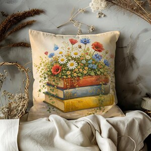 May include: A decorative throw pillow featuring a watercolor illustration of a stack of books topped with a vibrant floral arrangement. The flowers include red poppies, blue cornflowers, and white daisies. The pillow has a soft, neutral background.