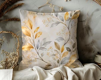 May include: Square decorative pillow with a botanical design. The pillow features a cream-colored background with a watercolor-style illustration of gold, gray, and brown leaves and small yellow flowers. The pillow is placed on a cream-colored fabric.