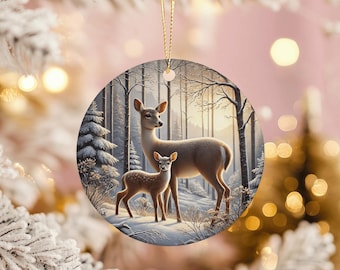 May include: A round Christmas ornament featuring a mother deer and fawn in a snowy forest scene. The deer are light brown with white accents, set against a backdrop of snow-covered trees and a soft, wintery light. A gold string is attached.