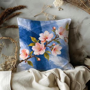 May include: A square throw pillow featuring a printed design of a cherry blossom branch with light pink flowers and green leaves set against a background of overlapping blue and white brushstrokes in a watercolor style.
