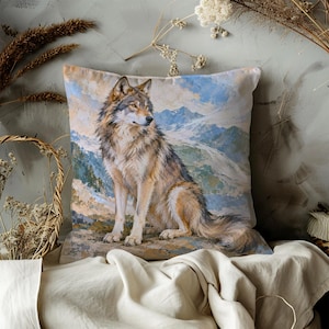 May include: A square throw pillow featuring a detailed illustration of a grey and brown wolf sitting on a rocky surface. The background depicts a painted landscape of blue mountains and a cloudy sky, rendered in a soft, artistic style.