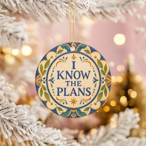 Puede incluir: Un adorno redondo de cerámica con el texto I KNOW THE PLANS en letras mayúsculas azules. El centro es de color crema, rodeado por un borde decorativo con motivos florales azules, verdes y naranjas. Cuelga de un hilo dorado fino.