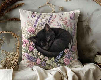 May include: Square throw pillow featuring a sleeping black cat surrounded by a floral wreath of pink, purple, and white flowers. Two butterflies are visible. The pillow is on a white background.