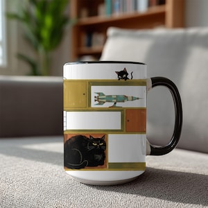 May include: White ceramic mug with a black handle and rim. The mug features a whimsical design of a black cat, a rocket ship, and other elements arranged in a shelf-like pattern. The mug is sitting on a gray surface.