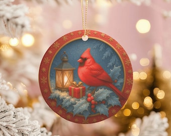 Peut inclure: Ornement de Noël rond représentant un cardinal rouge perché sur une branche de houx. L'ornement a une bordure rouge avec des motifs de flocons de neige et un fond bleu. Une lanterne et un coffret cadeau sont également représentés.