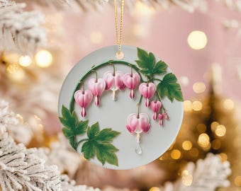 May include: A round Christmas ornament featuring a 3D depiction of pink bleeding heart flowers and green leaves. The ornament has a light gray background and a gold hanging string. The flowers are detailed with a realistic appearance.
