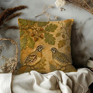 May include: A square throw pillow featuring a printed design of two quail birds standing among green oak leaves and brown acorns. The background has a textured, tan-coloured appearance with faint, script-like writing layered underneath the nature motif.
