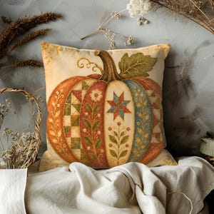 May include: A square throw cushion featuring a large central pumpkin illustration. The pumpkin is divided into vertical segments decorated with patchwork quilt patterns, floral vines, and a star motif in shades of orange, green, blue, and cream.