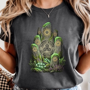 May include: Dark gray t-shirt featuring a graphic of standing stones with sun symbols and the text "Ancient Wisdom The Wheel Turns". The design includes green moss and purple flowers.