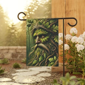 May include: A garden flag featuring a detailed illustration of a Green Man. The artwork depicts a face composed of leaves and foliage, with a long, flowing beard. The flag is displayed on a black metal stand, set against a blurred outdoor background.