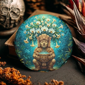 May include: Round, decorative art piece featuring a stylized stone figure holding a bowl with blue vapor. The figure is adorned with a floral headdress of white orchids and yellow trumpet flowers, set against a teal background with lush foliage.