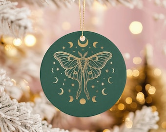 May include: A round, green Christmas ornament with a gold moth and crescent moon design. The ornament is hanging from a gold string. The background is a blurred image of a Christmas tree.