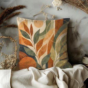 May include: Square decorative pillow with an abstract botanical design. The pillow features a pattern of stylized leaves in shades of orange, green, and beige, with geometric shapes. The pillow is placed on a white cloth.
