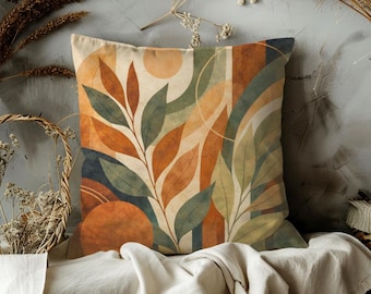 May include: Square decorative pillow with an abstract botanical design. The pillow features a pattern of stylized leaves in shades of orange, green, and beige, with geometric shapes. The pillow is placed on a white cloth.