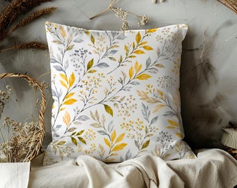 May include: Square throw pillow with a white background and a botanical pattern of yellow, gray, and green leaves and small flowers. The pillow is placed on a cream-colored fabric.