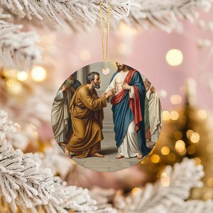May include: Round Christmas ornament featuring a religious painting of Jesus with disciples. The artwork depicts figures in robes, with a gold string for hanging. The background is a soft pink with bokeh lights and a frosted white tree branch.