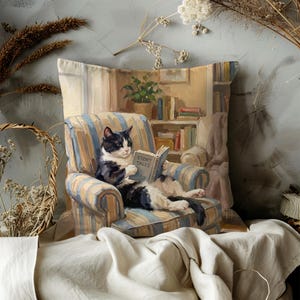 May include: Square decorative pillow featuring a black and white cat reading a book titled "Story Tales" while seated in a striped armchair. The pillow has a painted illustration style with a cozy, indoor scene.