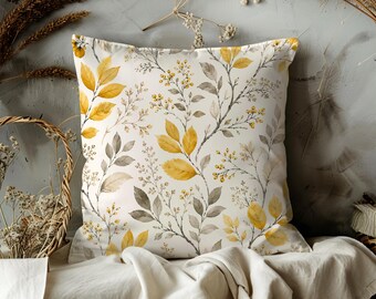 May include: Square throw pillow with a botanical design. The pillow features a white background with a pattern of yellow and gray leaves and small yellow berries. The pillow is placed on a white cloth.