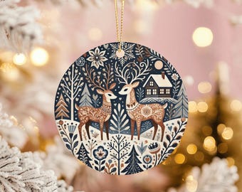 May include: Circular Christmas decoration depicting a winter scene with two deer, trees, and a house. The ornament has a dark blue backdrop with white, brown, and beige details. A gold string is attached for hanging.