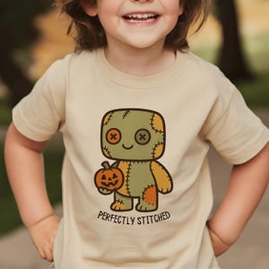 May include: A tan t-shirt featuring a cartoon character with a stitched-together design, holding a jack-o'-lantern. The character has a green head and body with orange patches and button eyes. The text "PERFECTLY STITCHED" is printed below the character.