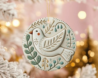 May include: A round, off-white ceramic ornament featuring a raised, embossed design of a bird surrounded by stylised botanical branches and leaves. The bird is accented with a small orange beak and a heart detail, while the foliage uses muted green and tan tones.