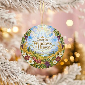 Puede incluir: Un adorno redondo de cerámica con una ilustración de paisaje vibrante con trigo, uvas y flores. El texto "Open the Windows of Heaven" y "Malachi 3:10" está impreso en dorado en el centro, bajo un cielo azul brillante con nubes.