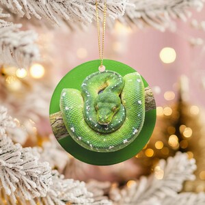 May include: Round Christmas ornament featuring a coiled green snake on a branch against a green background. The snake has white spots and is centered on the ornament. A gold string is attached for hanging. The background is blurred with bokeh lights.