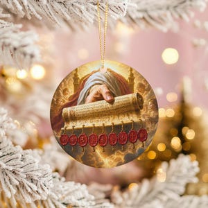 May include: Round Christmas ornament featuring a religious image. The ornament depicts a hand holding a scroll with text and red seals. The background has a golden glow with clouds and architectural elements. A gold string is attached for hanging.