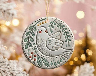 May include: A round, white ceramic Christmas ornament featuring a dove design. The dove is surrounded by green leaves and red berries. The ornament is suspended by a gold string.