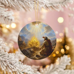 May include: Round Christmas ornament featuring a painting of a biblical scene. The artwork depicts a man kneeling before a burning bush, with a mountainous landscape and a radiant sky. The ornament is suspended by a gold string.