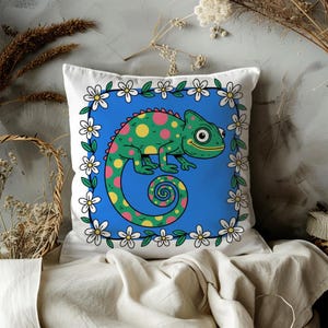 May include: White square throw pillow featuring a cartoon chameleon with green, yellow, and pink spots on a blue background. The pillow is framed with a border of white daisies and green leaves.