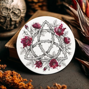 May include: A white round coaster featuring a black and white illustration of a Triquetra knot with thorny vines and burgundy roses. The coaster has a dark background with a skull and book in the background.