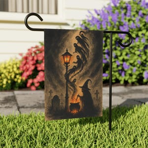 May include: A Halloween garden flag featuring a spooky scene with a witch stirring a cauldron, ghosts, a lamppost, and a graveyard. The flag is displayed on a black metal stand in a grassy yard, with colorful flowers in the background.