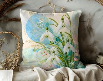 May include: Square decorative pillow with a floral design. The pillow features a watercolor painting of white snowdrop flowers with green stems and leaves against a light blue and cream background. The pillow is placed on a neutral-colored surface.