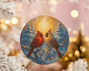 May include: Round Christmas ornament featuring a painting of two red cardinal birds perched on a branch. The background depicts a winter scene with blue trees and a yellow sun. The ornament has a gold string for hanging.