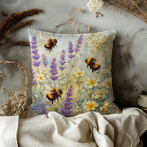 May include: Square decorative pillow featuring a watercolor design of bumblebees, lavender, and wildflowers. The pillow has a light blue background with yellow, purple, and white floral elements. The bees are black and yellow.